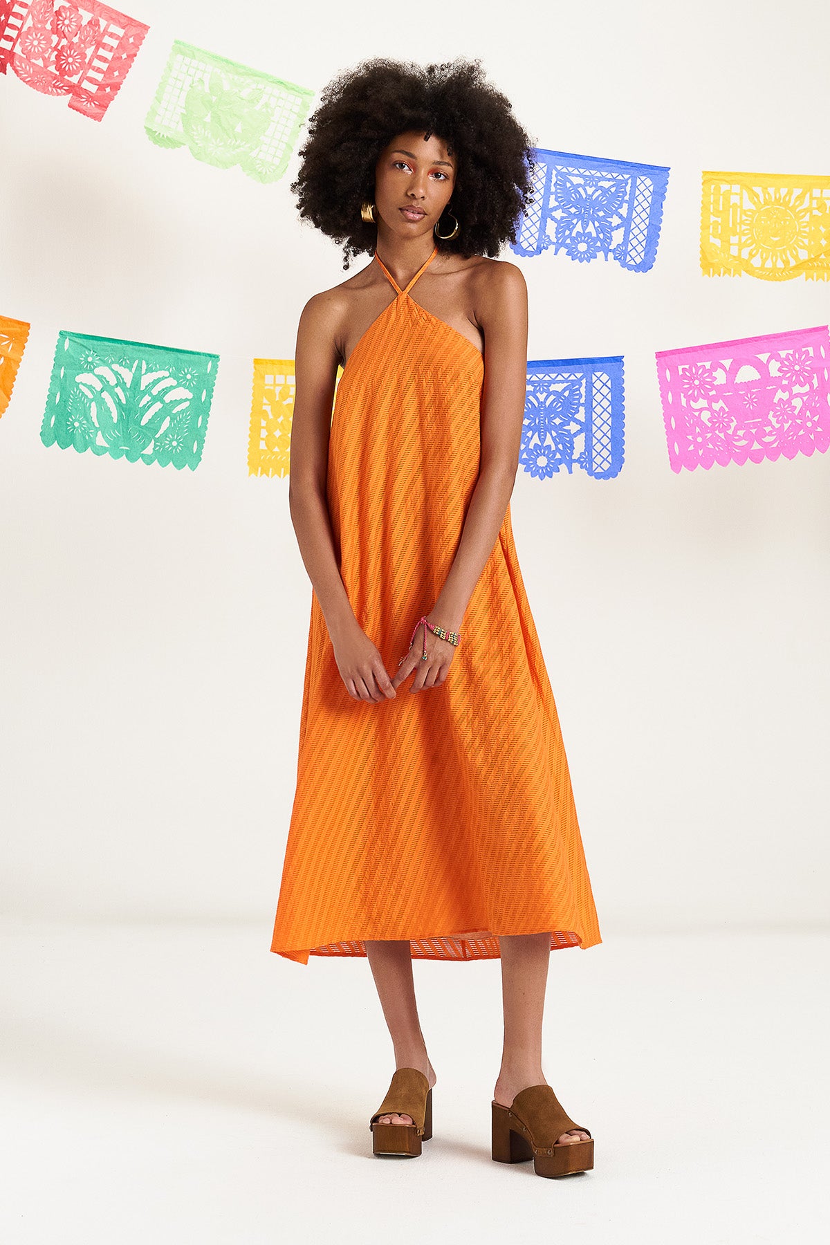 De Orangina 3/4 jurk in levendig oranje kleur en losse pasvorm, sluit achteraan in de hals met lintjes en zijn afgewerkt met speels detail van kwastjes en gouden pareltjes.