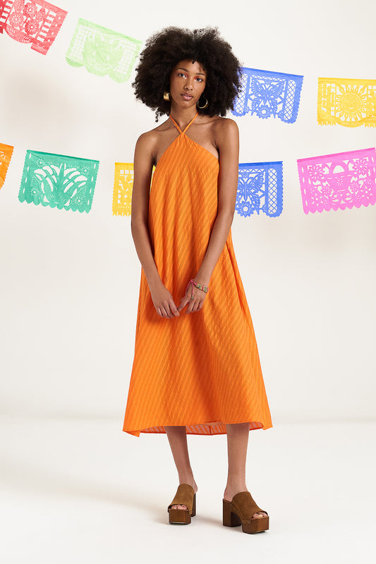De Orangina 3/4 jurk in levendig oranje kleur en losse pasvorm, sluit achteraan in de hals met lintjes en zijn afgewerkt met speels detail van kwastjes en gouden pareltjes.